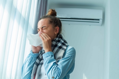 Woman standing in front of AC sneezing into tissue